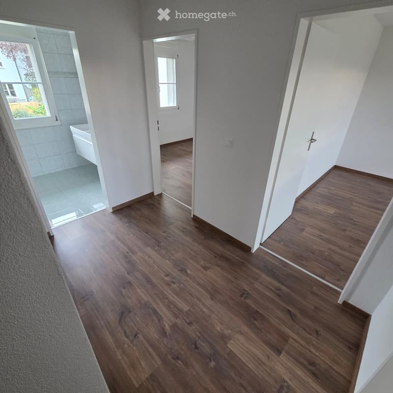 Empty hallway with wooden flooring, white walls, and doors leading to other rooms, including a bathroom with a bathtub visible through an open door.