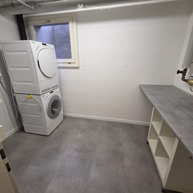 Small bathroom with a washing machine and dryer stacked, a shelf on the right side, and a window above the machines.