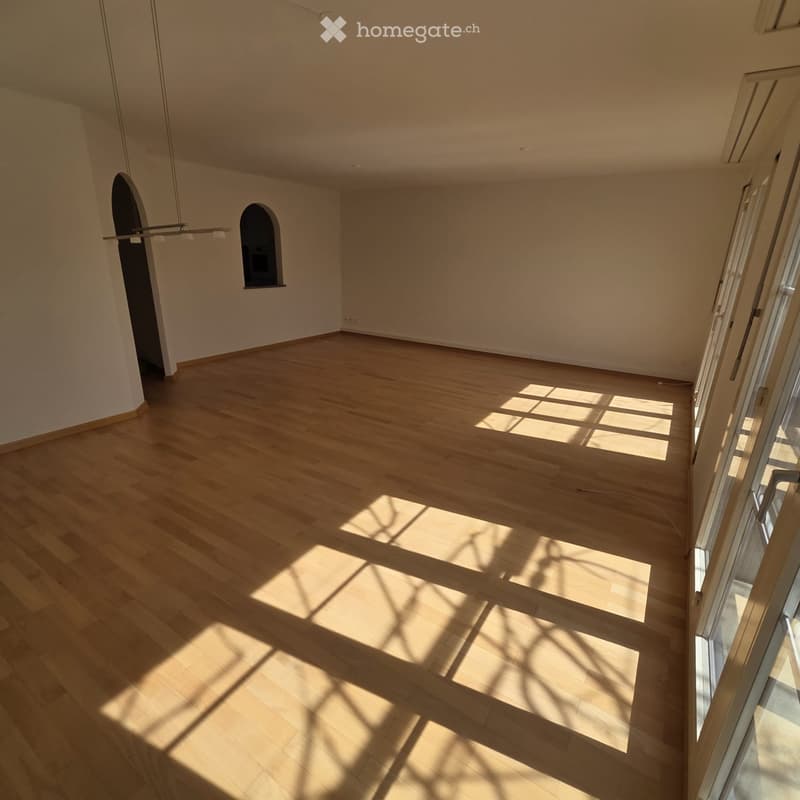 Empty room with light wooden flooring and white walls, featuring a large window with blinds and a small arched opening in the wall.