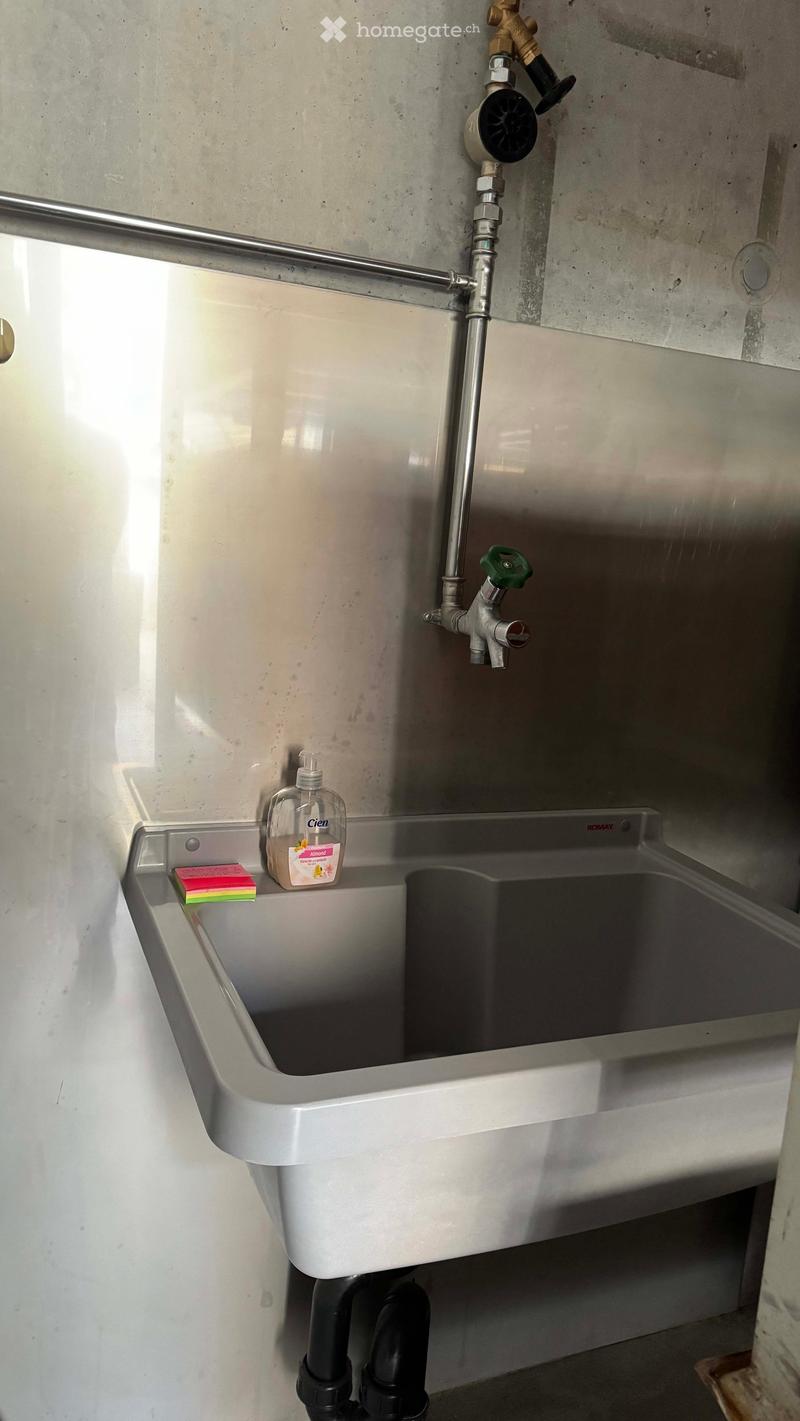 A stainless steel sink with a faucet, soap dispenser, and soap placed on the edge. The sink is mounted on a concrete wall with visible plumbing.