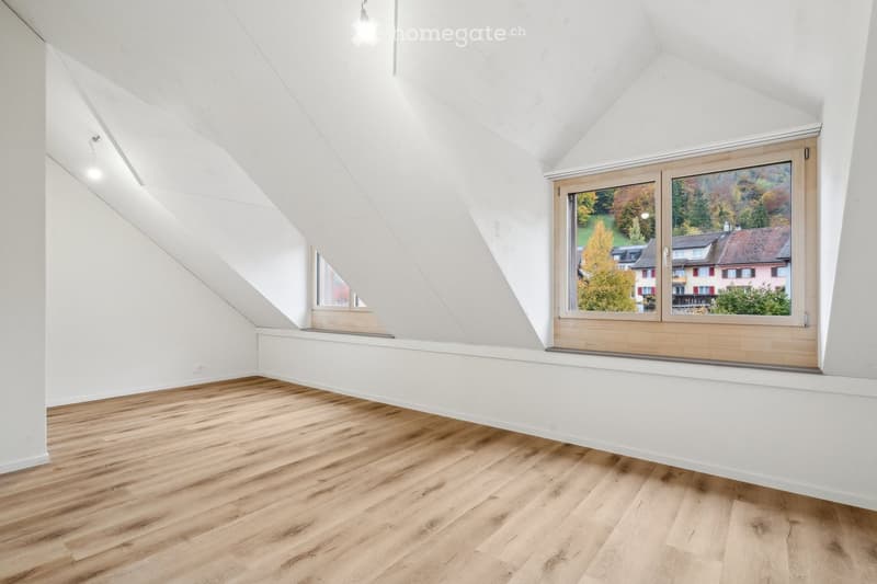 Empty attic room with wooden flooring, white walls, and large windows offering a view of the surrounding landscape.