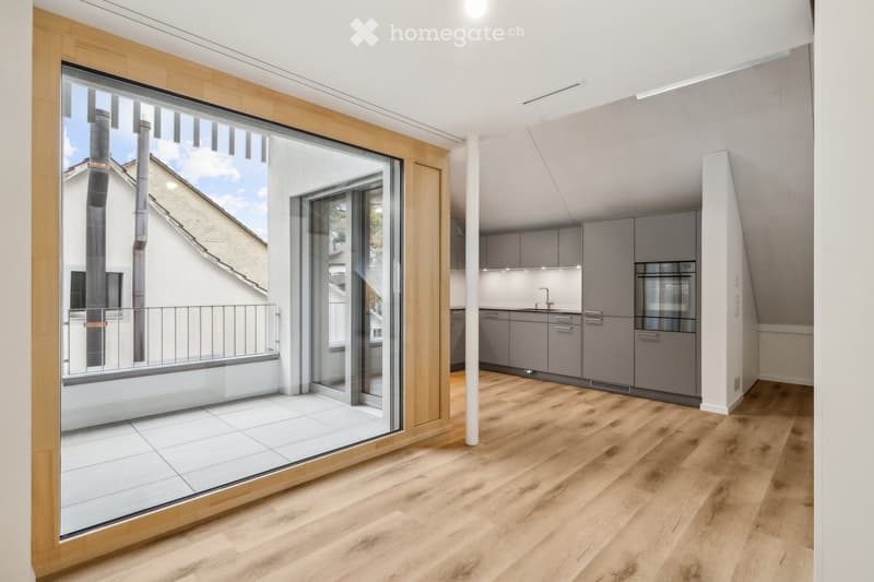 Modern, open-plan space with a balcony area and a kitchen. Features include wooden flooring, grey kitchen cabinets, a built-in oven, and a large sliding glass door leading to a tiled balcony.