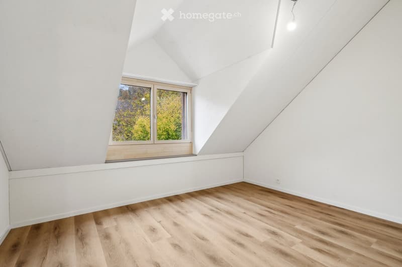 Empty attic room with wooden flooring, white walls, and a large window offering a view of trees.