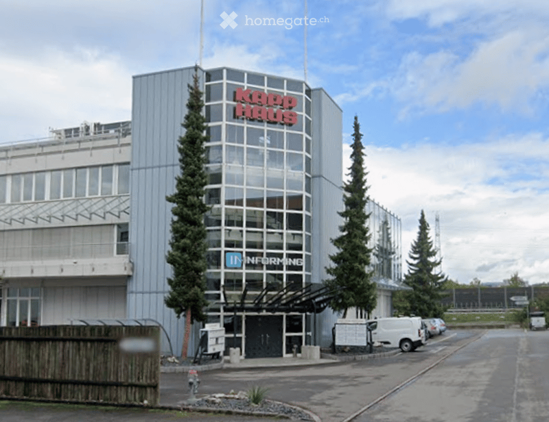 Modern multi-story building with large glass windows and a sign reading "KADO HOUSE" in red letters. The building has a sleek, contemporary design with a mix of metal and glass facades. There are several parked cars in front, and the surrounding area incl