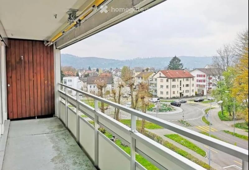 Balcony with metal railing, overlooking a town with houses, trees, and distant hills.