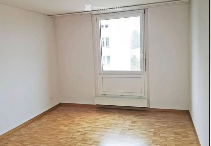 Empty room with wooden flooring, white walls, and a large window with white frame and radiator below.