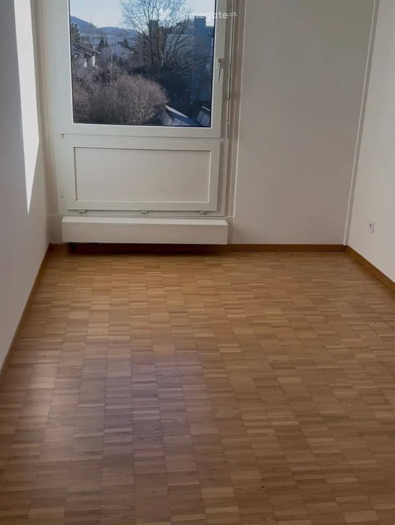 Empty room with wooden flooring, white walls, and a large window with a view of the outside.