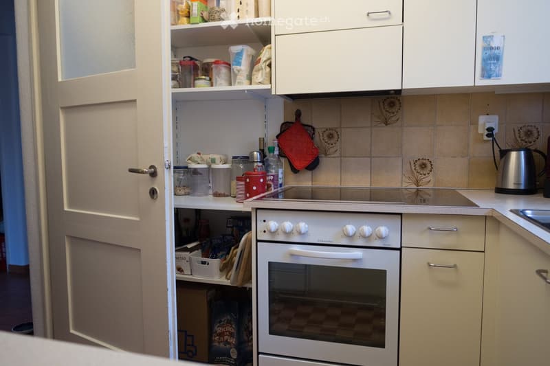 A compact kitchen with white cabinets, an oven, a shelf with food items, and a kettle on the counter.