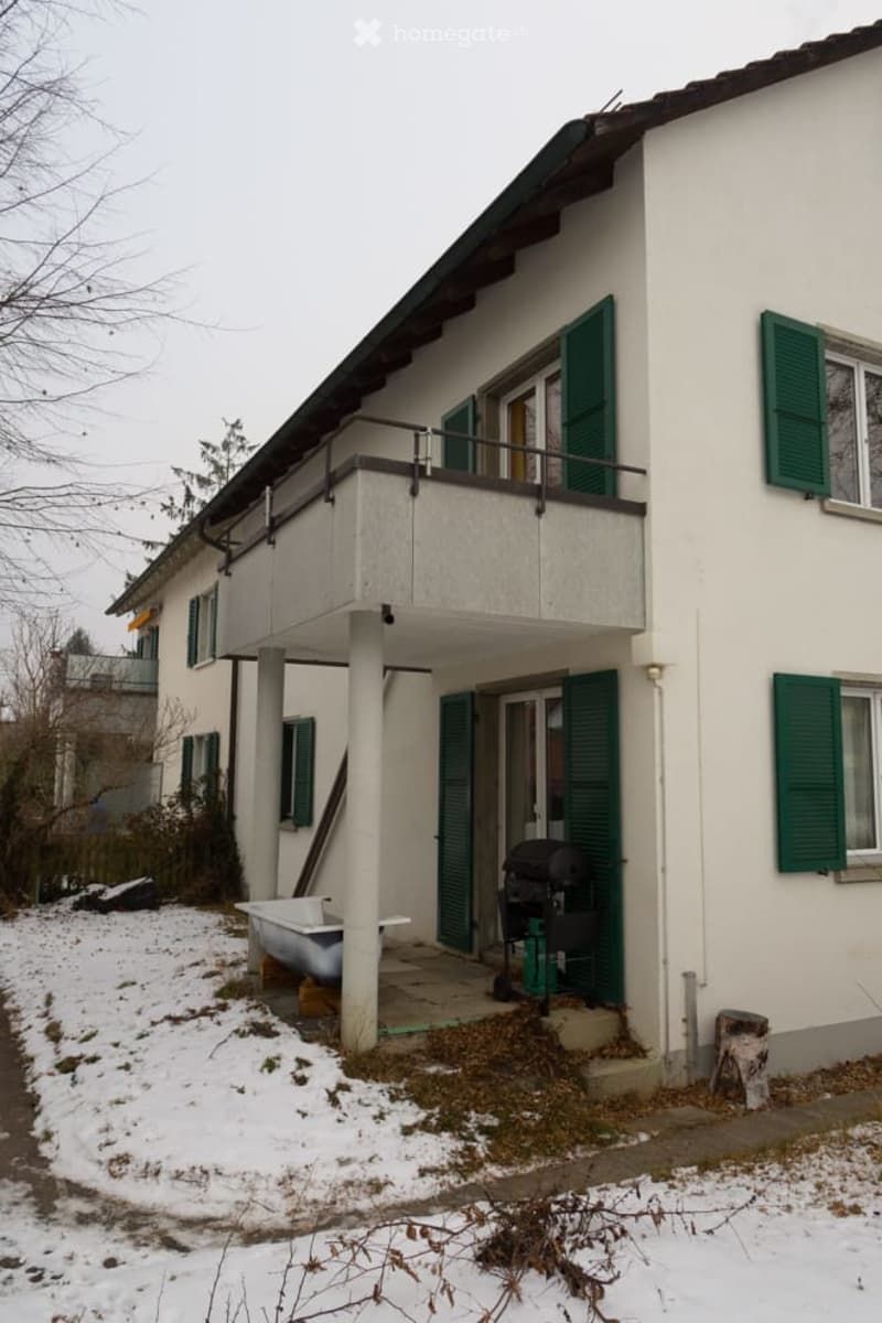 Two-story house with white exterior, green shutters, and a balcony on the upper floor. Snow is visible on the ground and roof.