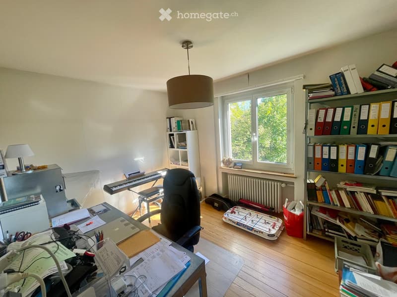 A well-lit office space with a large window, wooden flooring, and various work and storage items including a desk, chair, laptop, keyboard, bookshelf, filing cabinet, and a small football table.