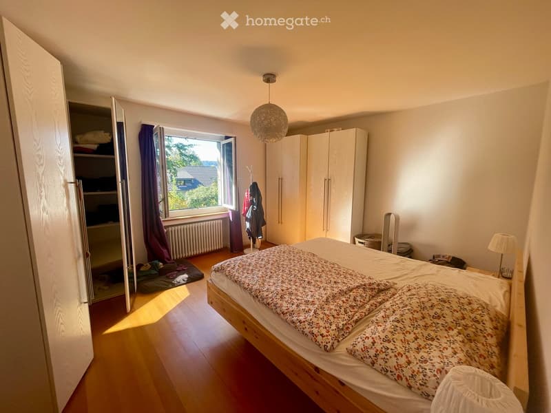 Cozy bedroom with wooden flooring, a large bed with floral bedding, a wardrobe, a hanging lamp, a window with purple curtains, and a coat rack with jackets.