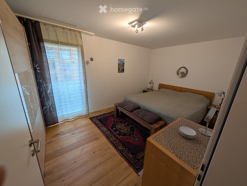 Cozy bedroom with wooden flooring, a large bed with pillows and a blanket, a nightstand with a lamp, a small table with a chair, a decorative rug, and artwork on the walls.