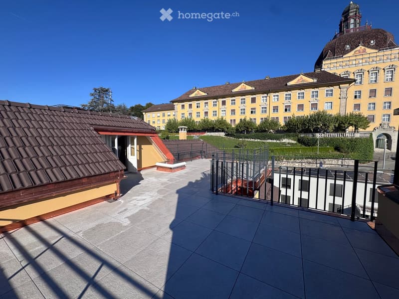 Roof terrace with tiled flooring, metal railing, and view of a large yellow building with a dome and multiple windows.