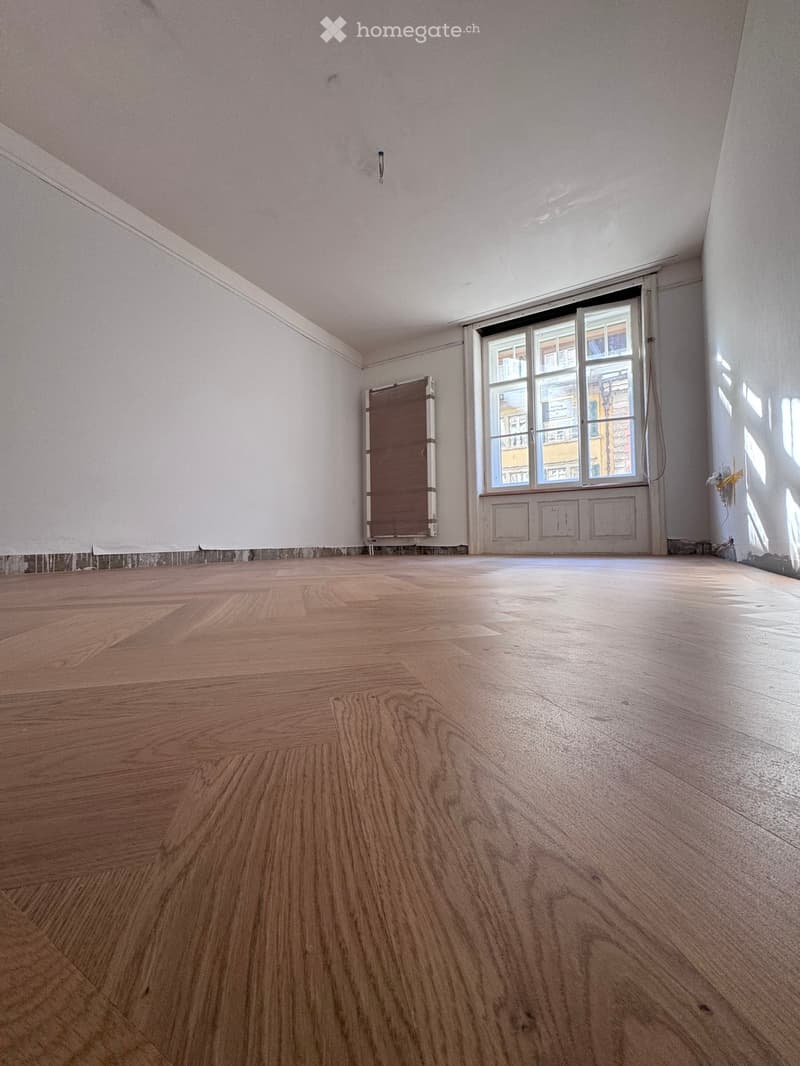 Empty room with wooden flooring, white walls, and a large window with a view of the street.