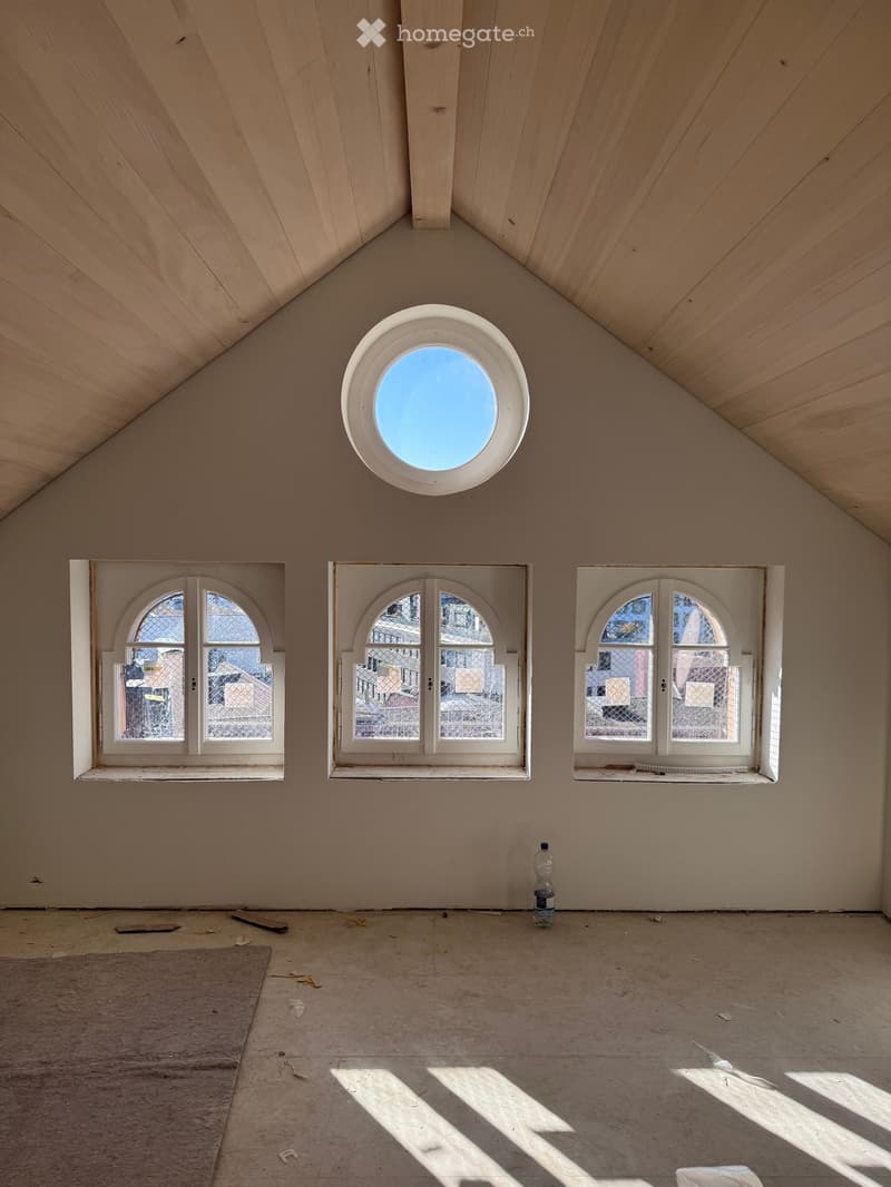 Empty attic room with wooden slanted ceilings, three arched windows, and a round skylight, showing natural light and construction materials on the floor.
