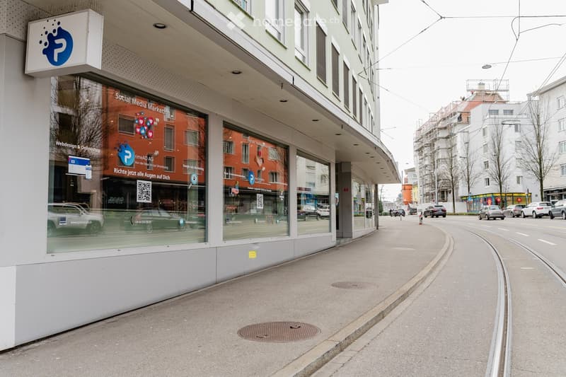 Large storefront with display windows, modern design, glass entrance, reflective surface, QR code on window, streetlight, tram tracks, parked cars, buildings, street