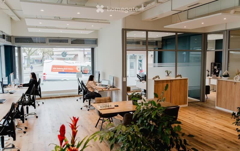 Spacious office space with large windows, wooden floors, ergonomic chairs, desks, monitor, and a game board on a table. Office furniture, potted plants, and equipment.