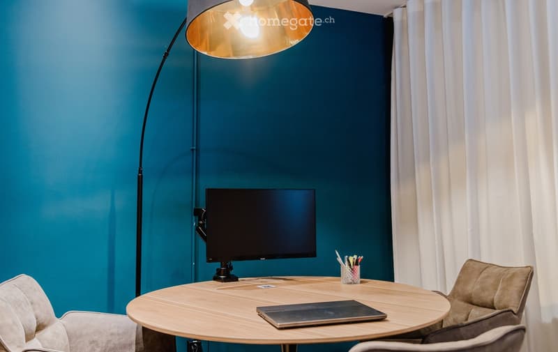 circular wooden table with monitor, pencil holder, laptop, wall lamp, blue wall, white curtain, armchairs