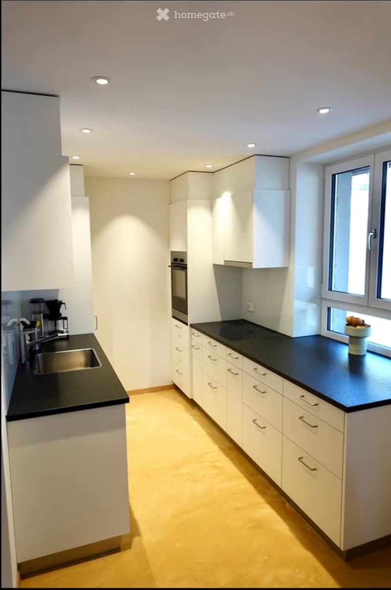 Modern kitchen with white cabinets, black countertops, and a stainless steel sink. Features include a coffee maker on the counter and a bowl of fruit near the window.