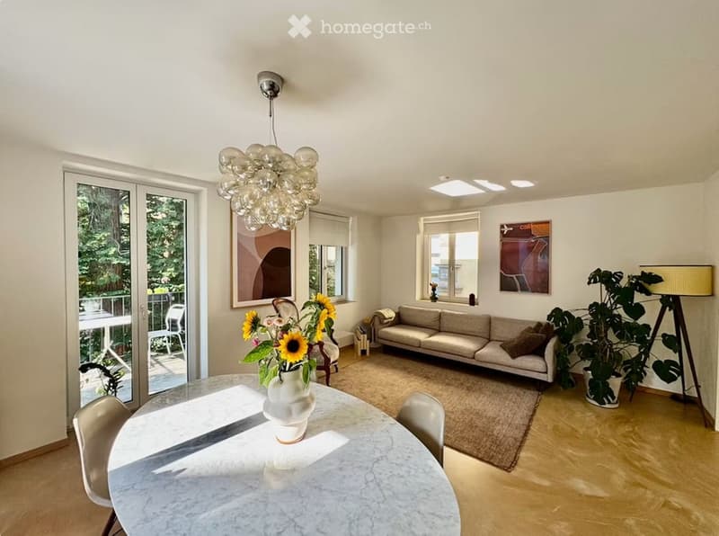 Bright living and dining area with a modern sofa, dining table with chairs, a large chandelier, and a view of a balcony.