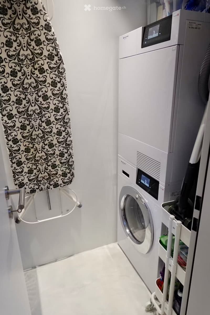 Modern bathroom with white tiled floor and walls, featuring a washing machine, dryer, and an ironing board with a patterned cover.