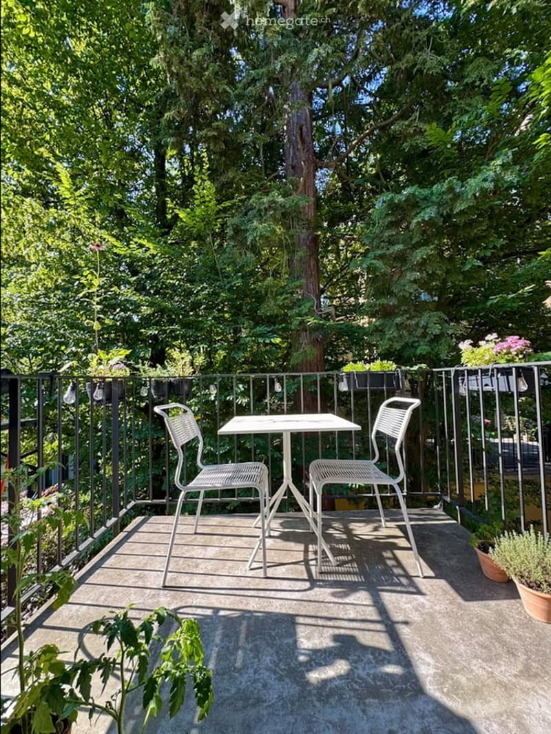 Balcony with a small table and two chairs, surrounded by lush greenery and potted plants.