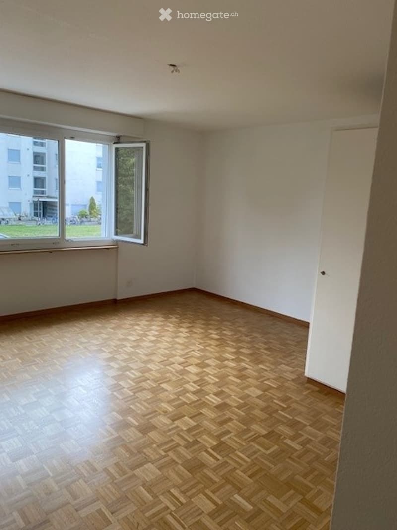 Empty room with wooden parquet flooring, white walls, and large windows with open shutters, allowing natural light to fill the space.