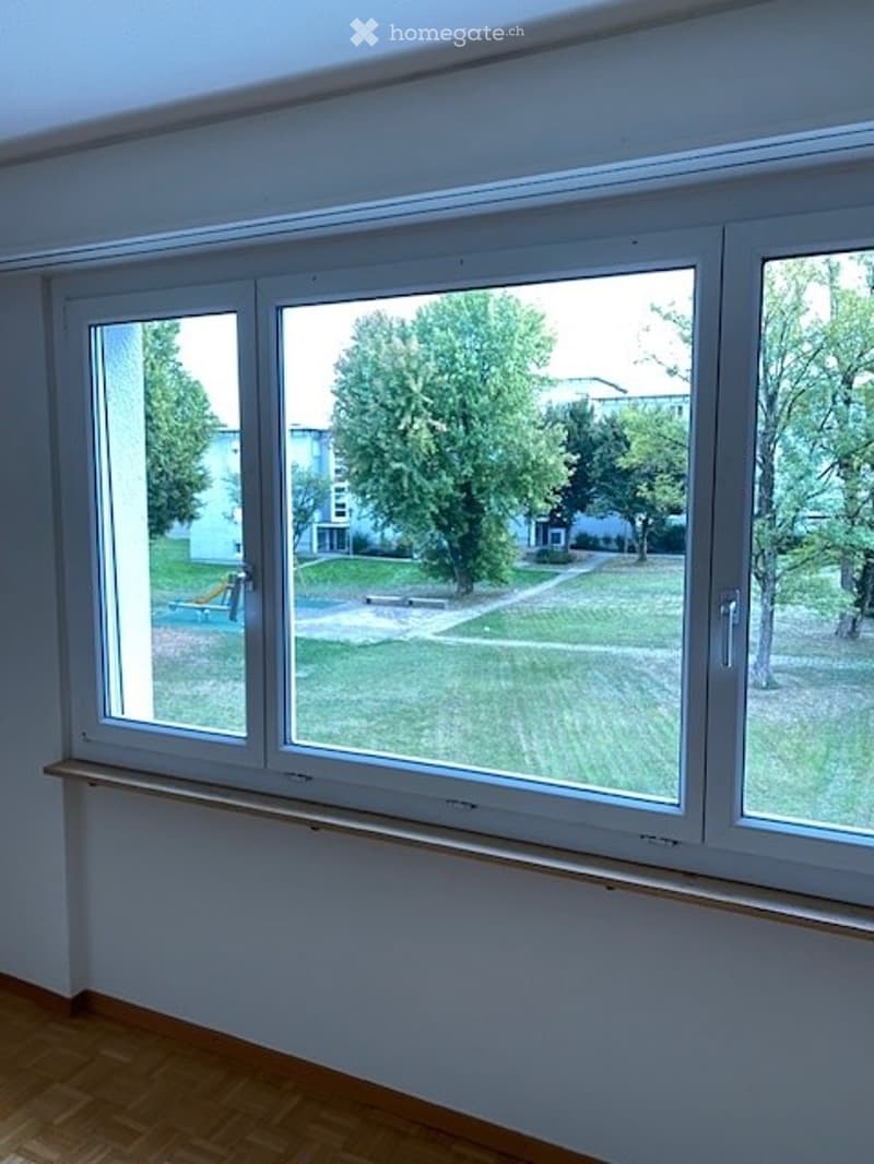 Empty room with wooden flooring and large windows showing a green outdoor area with trees and a playground.