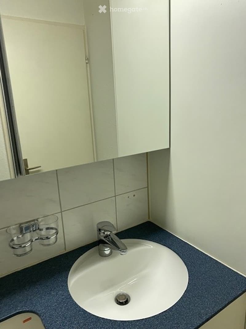 A compact bathroom with a white sink, chrome faucet, and a mirrored cabinet above it. The walls are tiled in light gray, and the countertop is a dark blue material.