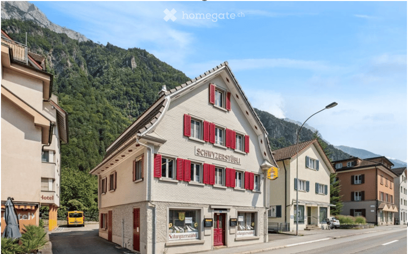 Traditional Swiss building with red shutters and signage reading "Schwyzenseeli" in a picturesque village setting with mountains in the background.