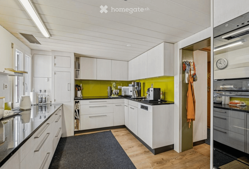 Modern kitchen with white cabinets, black countertops, and a green accent wall. Features include a microwave, coffee maker, oven, dishwasher, and refrigerator. Wooden flooring and a dark rug are present.