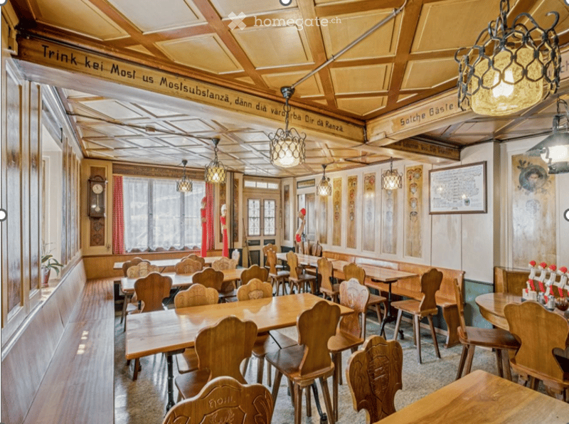Traditional restaurant interior with wooden tables, chairs, benches, and hanging pendant lights. Wooden ceiling beams and decorative signage are visible.