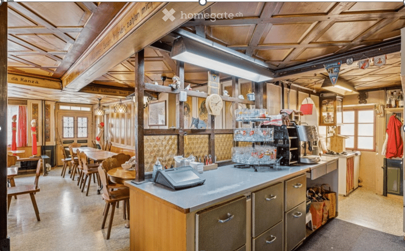 A rustic restaurant interior with wooden beams, a central counter, tables and chairs, and a cash register. The decor includes a clock, glassware, and various small items on shelves.