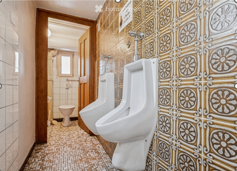 Public restroom with two urinals and a toilet, featuring patterned tiles and a wooden door leading to another room.