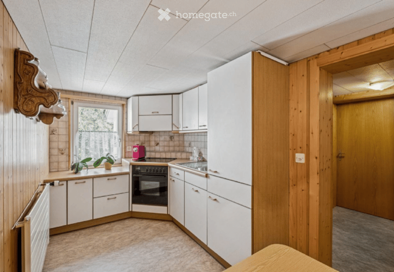 Bright kitchen with white cabinets, wooden accents, a central island, and a window overlooking a green area. Includes an oven, sink, faucet, radiator, and potted plants on the counter.