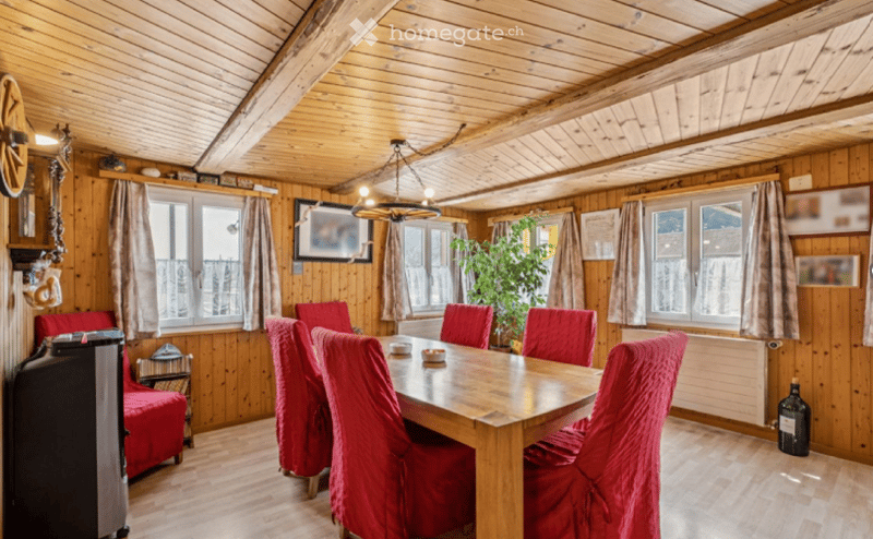 Cozy wooden-paneled dining room with a large wooden dining table surrounded by red chairs, a red sofa, a potted plant, and a chandelier hanging from the ceiling.