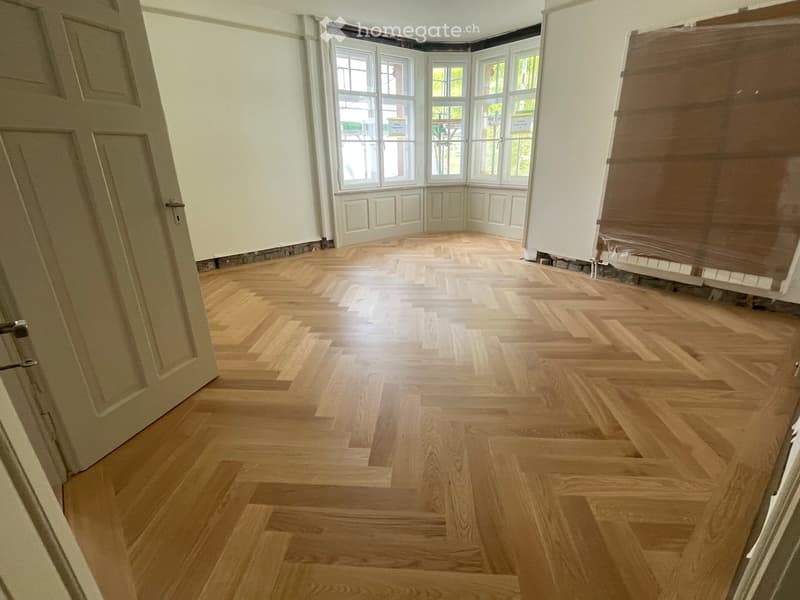 Empty room with light wooden herringbone flooring, white walls, and large windows with white frames. No furniture or equipment present.