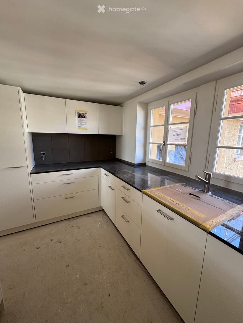 Empty kitchen with white cabinets, black countertops, and a sink area. Windows provide natural light, and the floor is unfinished concrete.
