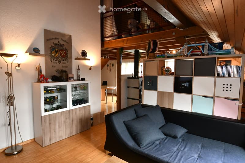 A modern living room with a black leather sofa, wooden floor, glass cabinet, shelves, and a floor lamp. The room features exposed wooden beams and a staircase leading to an upper level.