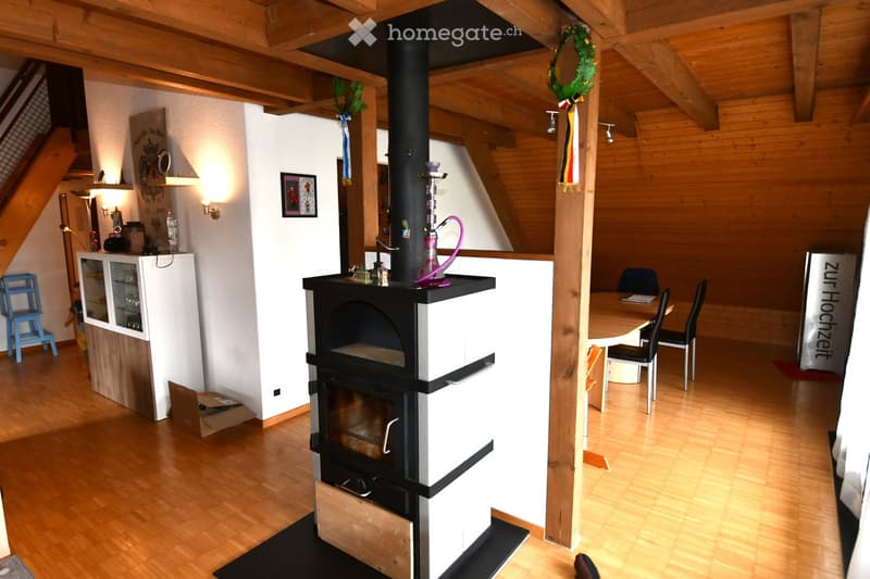 Cozy living room with exposed wooden beams, a central black wood-burning stove, a dining table with chairs, a small cabinet, and a blue stool near the staircase.
