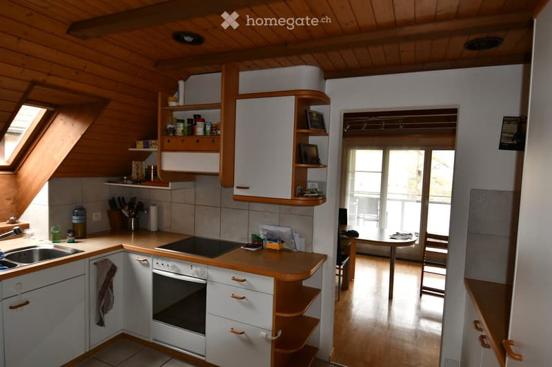 Modern kitchen with white cabinets, wooden accents, and built-in appliances. Features a sink, oven, and countertop with various kitchen items. A small dining area with a table and chairs is visible through an open doorway.