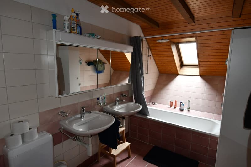 Bathroom with two sinks, a bathtub, a toilet, a mirror, and a window with wooden frame. The room features wooden ceiling beams and tiled walls.