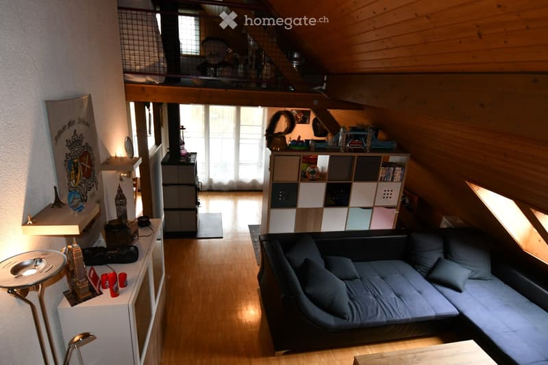 Cozy attic room with wooden beams, a black sofa, a small table, a lamp, a shelf with books and decorative items, and a mirror on the wall.