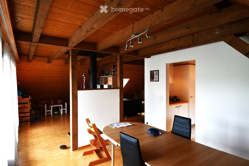 A spacious, well-lit room with wooden beams on the ceiling, a wooden dining table with chairs, a sofa, and a small cabinet. The floor is wooden, and there is a visible doorway leading to another area.
