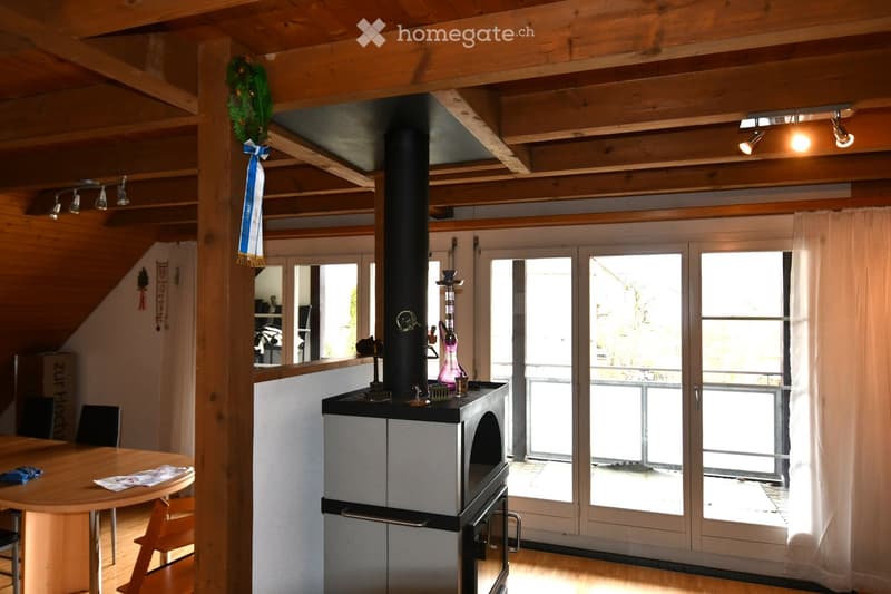 Cozy living room with wooden ceiling beams, a round wooden table with chairs, a modern fireplace, large windows with white curtains, and decorative items on the mantel.