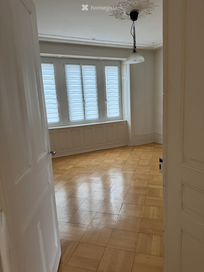 Empty room with wooden flooring, white walls, and large windows with shutters. A decorative ceiling light fixture is visible.
