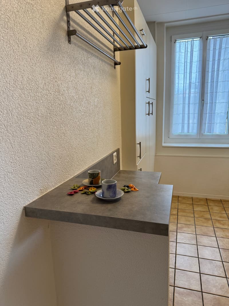 A small kitchen area with a countertop, two cups on plates, and colorful towels placed on the counter. The room has tiled flooring and a window with blinds.