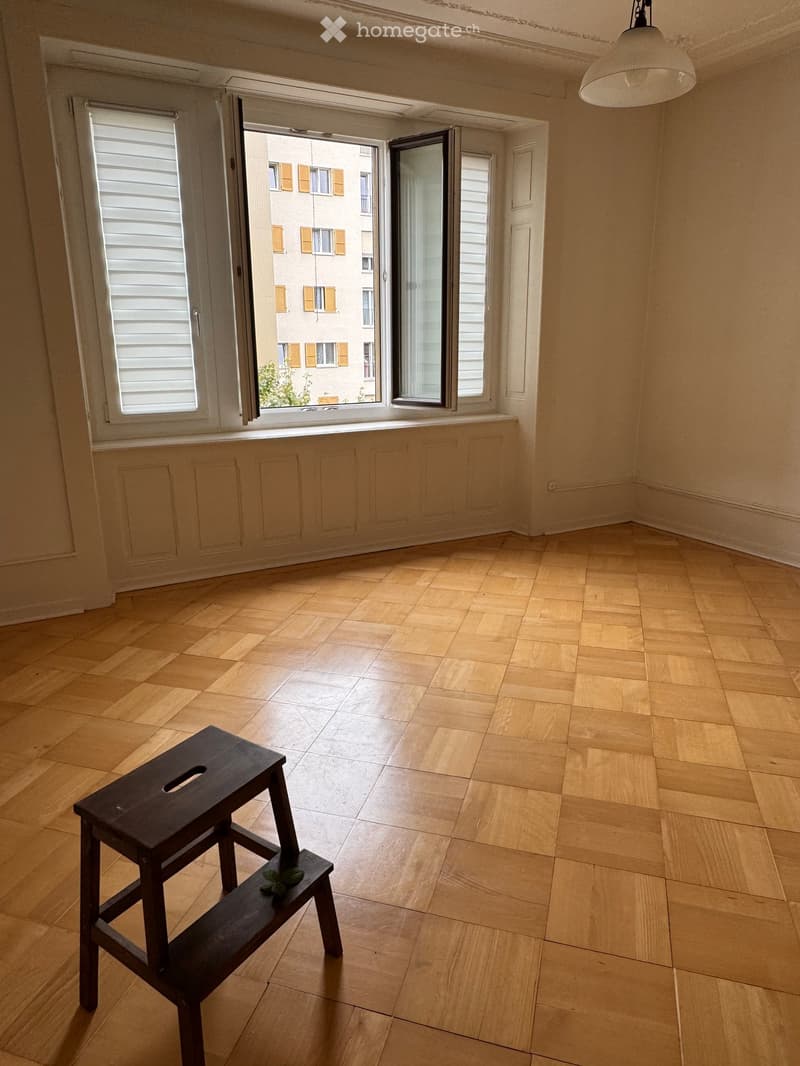 Empty room with wooden flooring and large windows, featuring a single dark wooden stool with a small plant on it.
