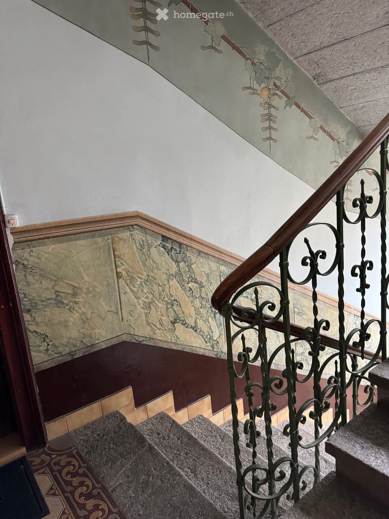 Staircase with ornate metal railing, marble wall, and patterned carpet leading upwards.