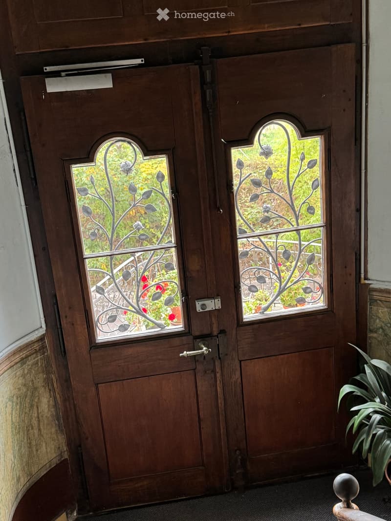 Wooden double doors with decorative glass panels featuring leaf designs, leading to a garden area with visible greenery and flowers.